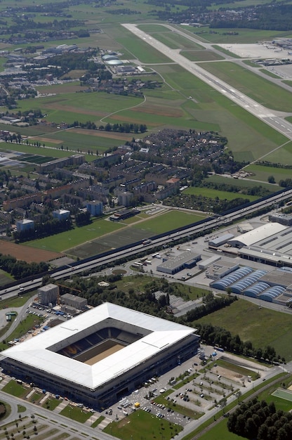 An aerial view of a sprawling automobile factory with a modern designed wing dedicated to electric vehicle battery production. Robotic arms are visible across the assembly lines, suggesting advanced automation. Steam or light haze rises from the plant, indicating industrial activity.