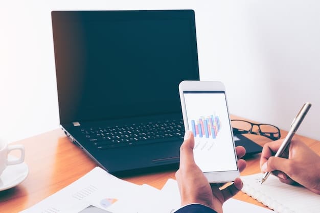 A person's hand holding a smartphone, displaying a financial tracking app with charts and graphs, while their other hand types on a laptop keyboard.