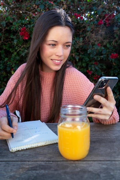 A college student with a laptop, looking at a budgeting app on their phone. There are healthy snacks and a reusable water bottle on the table, indicating mindful spending.